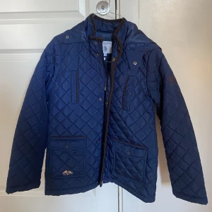 Blå quiltad jacka från USPA POLLO ASSN - Säljer en snygg blå quiltad jacka från Barbour. Den har långa ärmar och praktiska fickor framtill. Perfekt för höst och vår med sin stilrena design och bekväma passform. Jackan har både dragkedja och knappar för extra skydd mot kyla. Passar perfekt för en casual look!