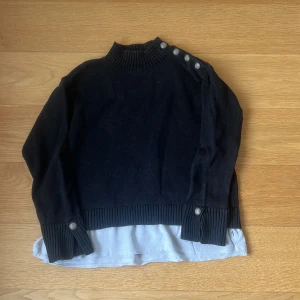 Svart stickad tröja från Rabalder - Säljer en svart stickad tröja från Rabalder i bra skick. Tröjan har en ribbad polokrage och knappar längs ena axeln och ärmsluten. Den har en vit underdel som ger en lager-på-lager-look. Perfekt för höst och vinter! Nypris 699kr 