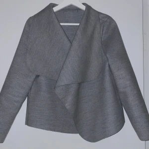 Grå kavaj liknande jacka med omlott-design - Säljer en stilren grå jacka med omlott-design. Perfekt för höst och vår, med långa ärmar och en elegant look. Passar både till vardags och festligare tillfällen. 