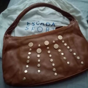 Brun axelväska från Escada - Säljer en snygg brun axelväska från Escada i skinn. Väskan har coola runda detaljer på framsidan och en bekväm axelrem. Perfekt för både vardag och fest! 👜