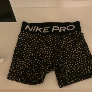 Svarta prickiga shorts från Nike Pro - Säljer ett par svarta Nike Pro shorts med guldiga prickar. De är perfekta för träning och har en tight passform. Shortsen är i mycket bra skick och har en elastisk midja för extra komfort. Perfekta för gymmet eller löprundan!❤️