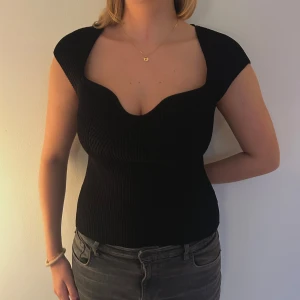 Svart ribbad topp - Snygg svart ribbad topp med en djup v-ringning. Den är tight och har korta ärmar, perfekt för både vardag och fest. Toppen är i ett stretchigt material som formar sig fint efter kroppen.