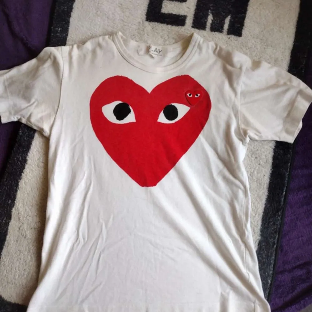 Säljer en vit t-shirt från Comme des Garçons med det ikoniska röda hjärtat med ögon på framsidan. Perfekt för en casual look eller att styla upp med en kavaj. T-shirten är i bra skick och gjord av mjuk bomull. Passar perfekt för både vår och sommar!. T-shirts.