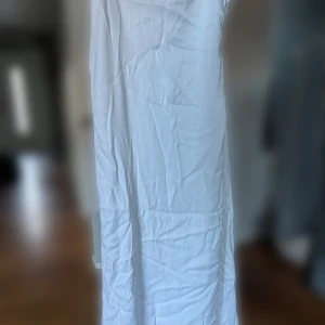 Vit klänning - Säljer en stilren vit klänning i lätt och bekvämt material. Perfekt för sommarens alla fester eller en avslappnad dag på stan. Klänningen har en elegant asymmetrisk design och är i bra skick. Har en liten öppning på sidan som går att justera i storlek.