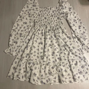 Blommig klänning  - Söt blommig klänning i vitt med små blå blommor. Den har långa ärmar och en smockad överdel som ger en fin passform. Perfekt för vår och sommar med volangdetaljer i nederkanten. Klänningen är i mycket bra skick, aldrig använt, inga hål eller fläckar. Säljs eftersom aldrig fick använda den 