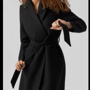 Svart kappa med bälte - Säljer en elegant svart kappa med bälte som ger en snygg siluett. Perfekt för höst och vinter. Den har långa ärmar och en klassisk krage. Passar både till vardags och festliga tillfällen. Ett måste i garderoben för den stilmedvetna!