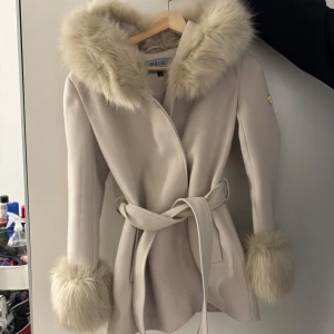 Beige kappa med päls från Hollies - Säljer en superfin beige kappa från Hollies i storlek 32. Den har en mysig fuskpäls på huvan och ärmsluten, perfekt för kyliga dagar. Kappan har ett bälte i midjan för en snygg passform. Den är i mycket bra skick (aldrig använd) och redo för höst och vinter! 
