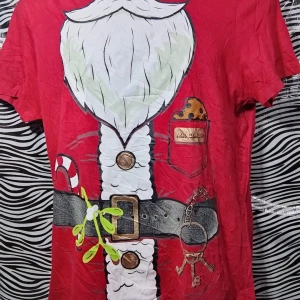 Röd jultröja med tomtemotiv - Rolig röd t-shirt med ett tryck av en tomtedräkt, komplett med skägg, bälte och godis. Perfekt för julfesten eller som en rolig present. Kortärmad och i bekvämt material.