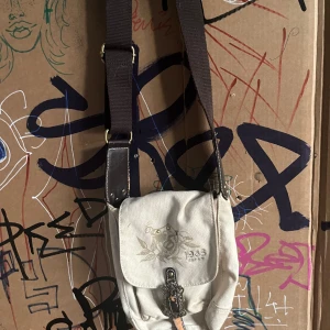 Beige axelväska emo - Säljer en beige axelväska med ett coolt tryck och texten '1968' på framsidan. Väskan har en justerbar brun axelrem och en dekorativ spänne på framsidan. Perfekt för vardagsbruk och ger en avslappnad stil. Den är i bra skick och redo för nya äventyr!