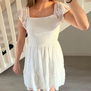 Vit sommarklänning / studentklänning - Säljer en superfin vit klänning perfekt för sommaren! Den har en smockad överdel som ger en bekväm passform och söta broderade blommor på ärmarna och kjolen. Klänningen är kort och har volangdetaljer. Perfekt för både vardag och fest! Perfekt till studenten, storleken passar en xsmall/small