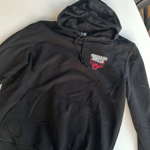 Svart Chicago Bulls hoodie från New Era - Säljer en svart hoodie från New Era med Chicago Bulls-logga i rött och vitt på bröstet. Den har en stor ficka fram och justerbar huva. Perfekt för basketfans eller bara för en chill stil. 🏀