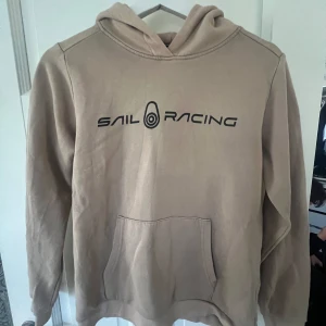 Beige hoodie från Sail Racing - Säljer en snygg beige hoodie från Sail Racing. Den har en klassisk känguruficka och en bekväm huva. Perfekt för en avslappnad stil och passar bra till både vardag och fritid. Loggan är tryckt i svart på bröstet. Skön och stilren design!
