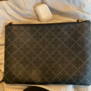 Svart kuvertväska med rutigt mönster - Snygg svart kuvertväska med diskret rutigt mönster. Väskan har en dragkedja upptill och är perfekt för en kväll ute eller en fest. Märket är malene birger, ny pris 900 kr 