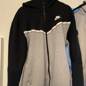 Svart och grå hoodie från Nike - Säljer en snygg och bekväm svart och grå hoodie från Nike i storlek L. Den har en dragkedja framtill och en klassisk Nike-logga på bröstet. Perfekt för höst och vår, eller bara för att chilla i. Huvan ger extra mysfaktor! 🖤