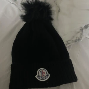 Svart stickad mössa från Moncler - Säljer nu min snygga svart stickade mössa från Moncler med en fluffig pälsboll på toppen. Den har en uppvikt kant och Monclers logga framtill. Aldrig använd, dock inget kvitto! 
