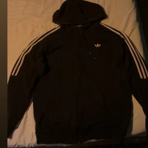 Svart huvtröja från Adidas - Säljer en svart huvtröja från Adidas i bra skick. Den har de klassiska tre vita ränderna längs ärmarna och en dragkedja framtill. Perfekt för höst och vår, och funkar både till träning och vardag. Storleken är normal och materialet känns mjukt och bekvämt.