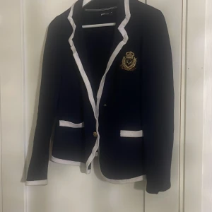 Mörkblå kavaj från Gina Tricot - Säljer en snygg mörkblå kavaj från Gina Tricot i storlek M. Den har vita kanter och ett elegant emblem på bröstet. Perfekt för både vardag och fest! Kavajen har en klassisk passform med knappar framtill. Passar perfekt till höst och vår.