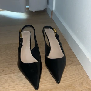 Svarta pumps med spetsig tå - Snygga svarta pumps i syntetmaterial med spetsig tå och elegant klack. De har en stilren design med en justerbar rem runt hälen. Perfekta för festliga tillfällen eller en utekväll. Skorna är i mycket bra skick och redo att användas för att lyfta din outfit!