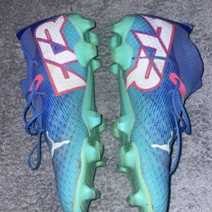 Blå fotbollsskor från Puma - Säljer ett par blå fotbollsskor från Puma i bra skick. Skorna har en cool design med vita och röda detaljer samt snörning för perfekt passform. Sulan är grön och anpassad för bra grepp på planen. Perfekta för fotbollsträning eller match!