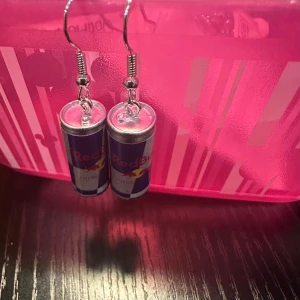 Örhängen med Red Bull-burkar - Unika örhängen formade som små Red Bull-burkar. Perfekta för att ge din outfit en rolig och energisk touch. De är silverfärgade med detaljer i blått och rött, och hänger från en enkel krok. Passar perfekt för fester eller när du vill sticka ut lite extra!