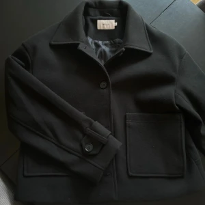 Svart kappa från Nelly - Säljer en stilren svart kappa från Nelly i storlek XS. Den har en klassisk design med knappar framtill och stora fickor. Perfekt för höst och vår, och ger en elegant touch till vilken outfit som helst. Passar både till vardags och festliga tillfällen.