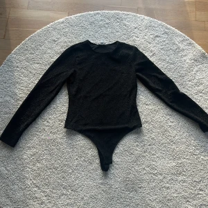 Svart glittrig body - Snygg svart body med glittrigt material som ger en festlig känsla. Den har långa ärmar och en tight passform som framhäver figuren. Perfekt för en utekväll eller fest! Bodysuiten har en enkel design med rund hals och knäppning i grenen.