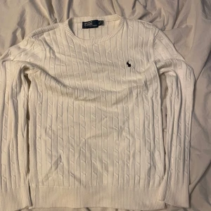 Vit stickad tröja från Polo Ralph Lauren - Säljer en snygg vit stickad tröja från Polo Ralph Lauren. Den har ett klassiskt kabelstickat mönster och en liten broderad logga på bröstet. Perfekt för både höst och vinter, 