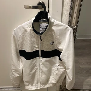Vit och svart jacka från Sergio Tacchini - Säljer en snygg vit och svart jacka från Sergio Tacchini i storlek XS. Jackan har en dragkedja framtill och ett stilrent mönster med en svart rand över bröstet. Perfekt för en sportig look eller vardagsstil. Passar som medium med 