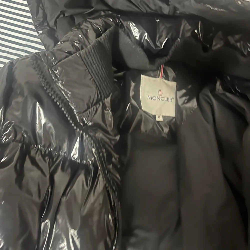 Säljer en snygg svart pufferjacka från Moncler i mycket bra skick. Jackan är perfekt för vintern med sitt blanka polyester-material och har en dragkedja framtill. Den har en cool serietidningsdetalj på insidan och är långärmad med en huva för extra värme. Passar perfekt för kalla dagar!. Jackor.