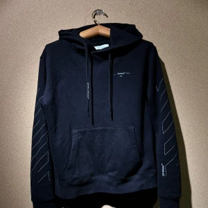 Svart OW hoodie - Oäkta snygg "unfinished" svart hoodie från OW med ikoniska vita pilar på ryggen och ärmarna. Den har en stor ficka fram och justerbar huva med snören. Perfekt för en casual look eller streetwear-stil. Passar både höst och vinter!