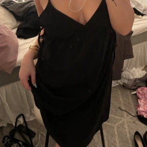 Svart klänning med knytband - Säljer en elegant svart klänning med knytband i sidan. Den har en djup v-ringning och är ärmlös, perfekt för festliga tillfällen. Klänningen är i bra skick och passar utmärkt för både sommar och nyår.