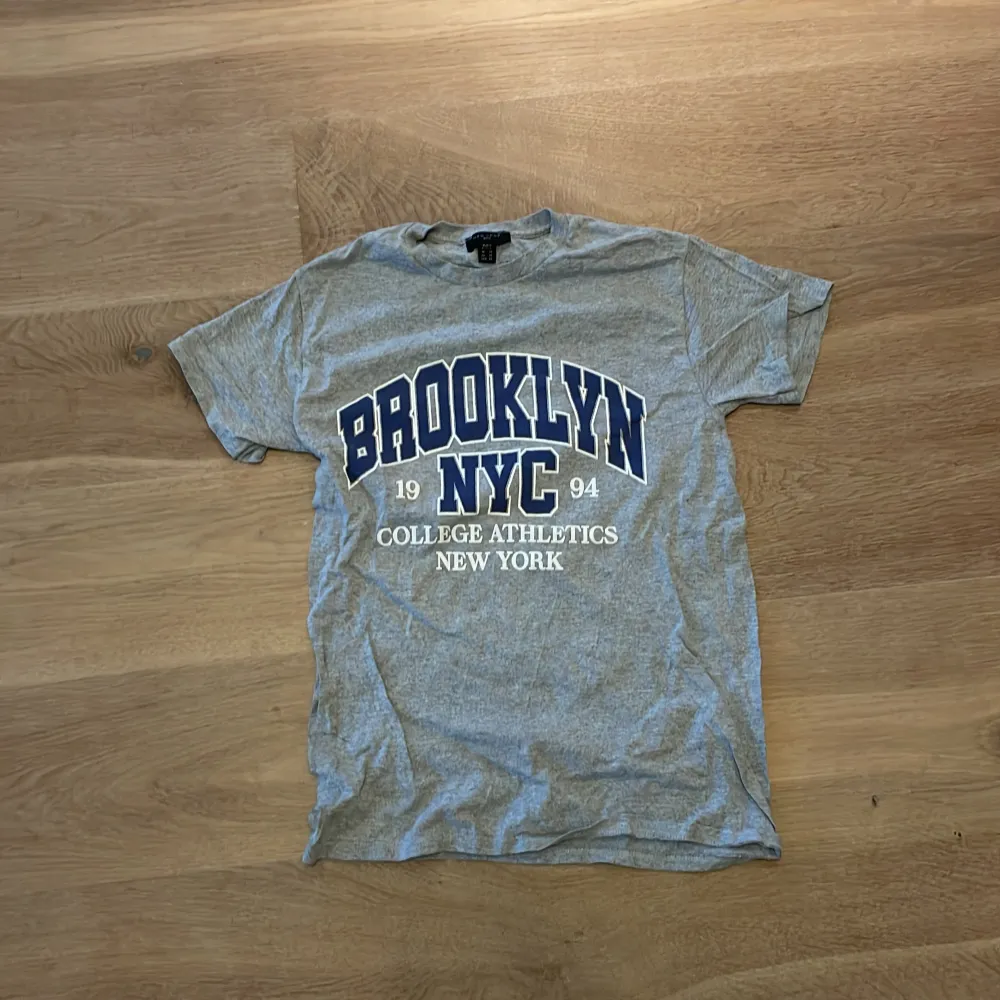 Säljer en grå t-shirt med stort tryck som säger 'Brooklyn NYC 1994 College Athletics New York'. Perfekt för en avslappnad stil, kanske till vardags eller träning. Den är kortärmad och i bra skick. Passar perfekt till jeans eller shorts!. T-shirts.