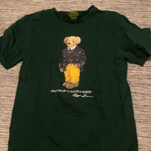 Grön t-shirt med björntryck från Ralph Lauren - Säljer en grön t-shirt från Ralph Lauren med ett coolt björntryck på framsidan. T-shirten har en klassisk passform och är perfekt för en avslappnad stil. Den är tillverkad i mjukt material för extra komfort. aldrig använd!!! pris kan diskuteras