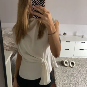 Vit knytblus från Zara - Säljer en stilren vit blus från Zara med knytning i midjan. Blusen har en elegant design med korta ärmar och en dragkedja i ryggen. 