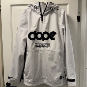 Vit jacka från Dope - Säljer en stilren vit jacka från Dope med svart logga på bröstet och ärmen. Jackan har en huva och dragkedja på sidan för enkel på- och avtagning. Perfekt för en sportig look.