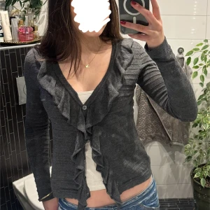 Grå kofta med volangdetaljer - Snygg grå kofta med volangdetaljer framtill och knappar. Den har en ribbad struktur och långa ärmar, perfekt för en stilren look. Passar bra till både jeans och kjol. Den är lite glittrig💕Passar både xs/s! Skriv för fler bilder☺️