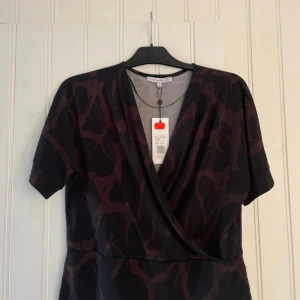 Brun omlottblus från Flash Woman - Snygg brun omlottblus från Flash Woman med ett unikt giraffmönster. Blusen har korta ärmar och en v-ringad design som ger en stilren look. Perfekt för att sticka ut med ett djärvt mönster.