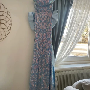 Blå långklänning med mönster - Elegant blå långklänning med ett vackert blommigt mönster i rosa och guld. Klänningen har en stor rosett baktill (den är avtagbar) och korta ärmar. Perfekt för speciella tillfällen.