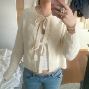 Vit stickad kofta med knytband - Säljer en mysig vit stickad kofta med knytband framtill. Koftan har långa ärmar och en ribbad struktur som ger en stilren look. Perfekt för att hålla sig varm och snygg under kyligare dagar. Passar bra till jeans eller kjol för en avslappnad stil.