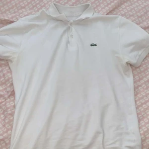 Vit pikétröja från Lacoste - Säljer en klassisk vit pikétröja från Lacoste med korta ärmar och den ikoniska krokodilloggan på bröstet. Perfekt för en stilren och avslappnad look. Tröjan har en knappslå med två knappar.