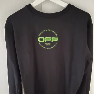 Svart tröja från Off-White - Snygg svart tröja från Off-White med ett unikt tryck av en hand och texten 'OFF' i grönt. Tröjan har långa ärmar och en rund halsringning. Perfekt för dig som vill ha något stilrent men med en twist.