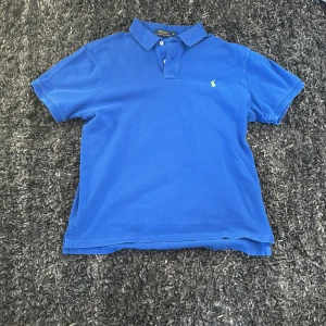Blå pikétröja från Ralph Lauren - Säljer en snygg blå pikétröja från Ralph Lauren i slim fit. Tröjan har korta ärmar och en klassisk krage med knappar. Den ikoniska loggan är broderad i grönt på bröstet. Perfekt för en stilren look!