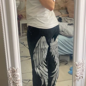 Svarta byxor med vingmönster -  Storlek: baggy L Coola svarta byxor med stort vitt vingmönster längs benen. Byxorna har en avslappnad passform och är perfekta för en unik stil. Märket 'Fashion Classicwear' är tryckt på framsidan.