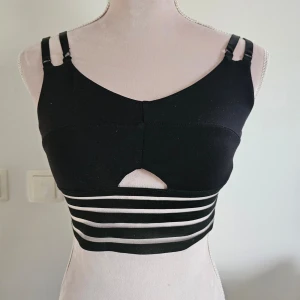 Svart sporttopp från Bombshell - Snygg svart sporttopp från Bombshell med dubbla axelband och randig detalj i midjan. Tillverkad i nylon och spandex för en bekväm passform. Perfekt för träning eller en aktiv dag. 