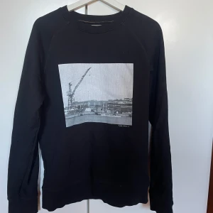 Svart tröja med tryck från Nudie Jeans - Säljer en svart tröja från Nudie Jeans med ett stort tryck av en kran och hamnlandskap på framsidan. Tröjan är långärmad och tillverkad av 100% ekologisk bomull. Perfekt för en stilren och avslappnad look. Knappt använd, storlek M