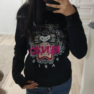 Svart tröja med tigertryck från Kenzo - Snygg svart tröja från Kenzo med ett iögonfallande tigertryck och texten 'Kenzo Paris' i rosa och vita detaljer. Tröjan har långa ärmar och en normal passform, perfekt för en trendig look.💗