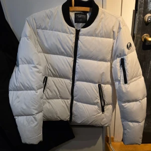 Vit pufferjacka från Peak Performance - Säljer en snygg vit dunjacka från Peak Performance. Jackan har en dragkedja framtill och praktiska fickor med dragkedjor. Den har en rund logotyp på ärmen och är perfekt för kyligare dagar. Jackan är långärmad och har en stilren design.