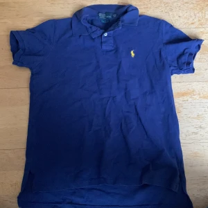 Blå pikétröja från Ralph Lauren - Säljer en klassisk blå pikétröja från Ralph Lauren med den ikoniska gula broderade loggan på bröstet. Tröjan har korta ärmar och en stilren krage med knappar. Perfekt för en avslappnad men stilren look. Obs passar S och XS