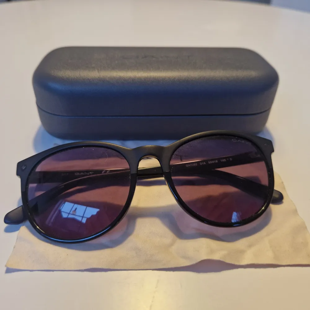 Snygga svarta solglasögon från Gant med runda glas och mörka linser. Kommer med ett praktiskt fodral och putsduk. Perfekta för en stilren look.. Asusteet.
