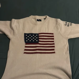 Vit stickad tröja från GANT med amerikansk flagga - Säljer en vit stickad tröja från GANT med en stor amerikansk flagga på framsidan. Tröjan har långa ärmar och texten 'GANT NH-CT 1949' på ena ärmen. Perfekt för en stilren och avslappnad look. Storlek s